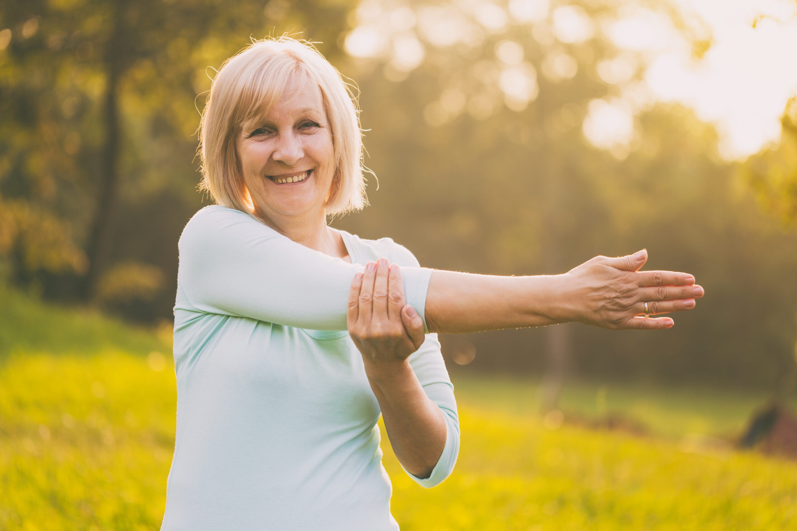 Smiling older woman stretching her arm outdoors in a sunlit park